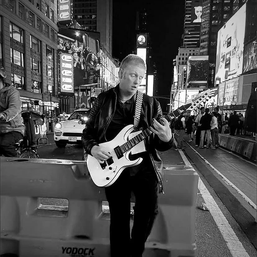Studio Raconteur - Cut Through The Noise Black and white photo of a man playing electric guitar in the middle of Times Square, New York, "cutting through the noise."